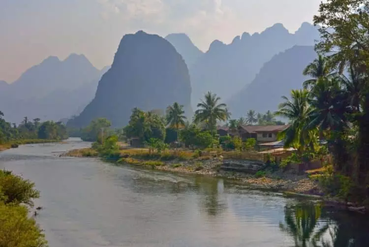 Village laotien bordant une rivière avec palmiers et montagnes karstiques en arrière-plan.