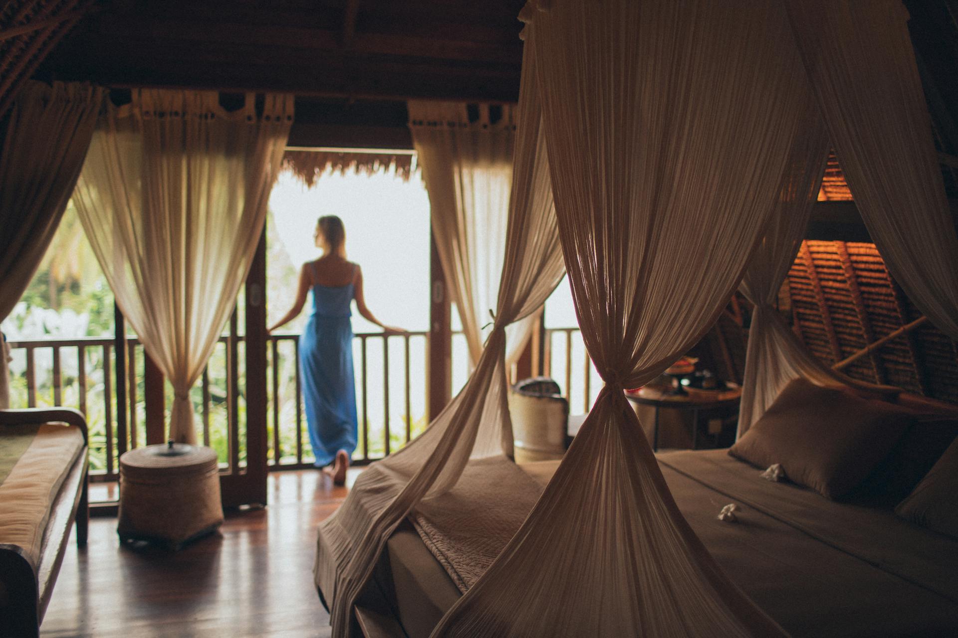 Femme en robe bleue contemplant un paysage depuis une chambre exotique.