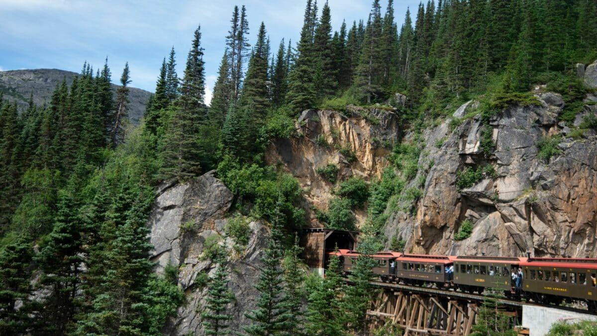 Train traversant une forêt dense et des falaises rocheuses en Alaska.