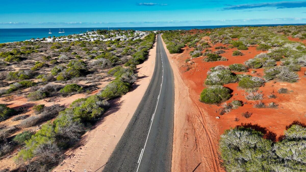 Route traversant un paysage aride australien avec sol rouge, végétation basse et océan en arrière-plan.