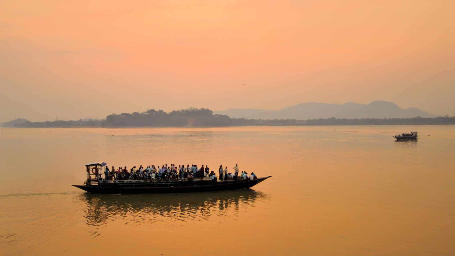 Bateau traditionnel naviguant sur le fleuve Brahmapoutre au coucher du soleil dans l’Assam, avec des passagers à bord.