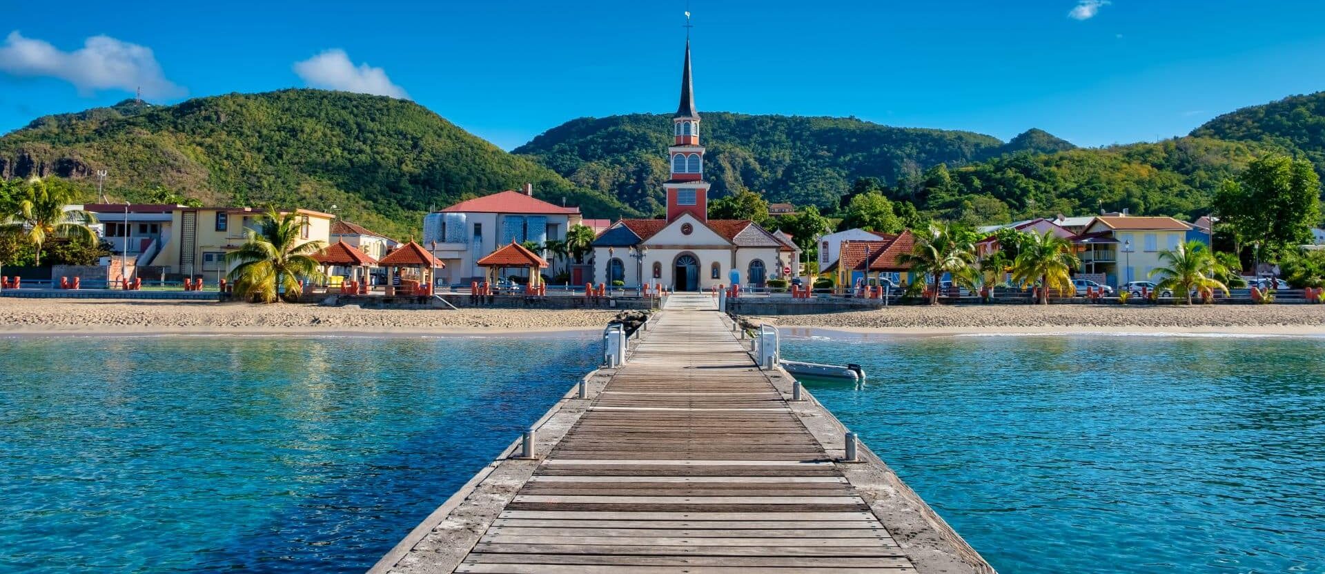 Village côtier de Martinique avec ponton en bois menant à une église face à la mer turquoise et collines verdoyantes en arrière-plan.
