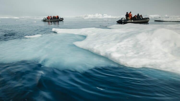Zodiacs avec passagers en combinaison rouge naviguant le long d’une plaque de banquise bleutée au Spitzberg.