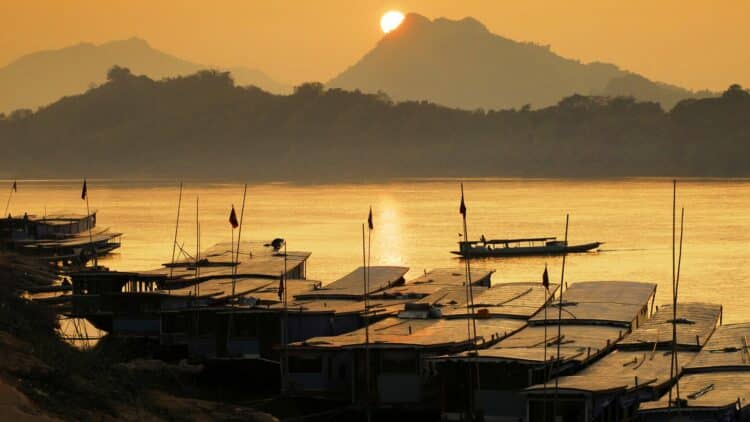 Bateaux traditionnels amarrés sur le Mékong au coucher du soleil avec montagnes en arrière-plan en Thaïlande.