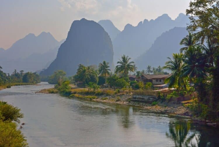 Village laotien bordant une rivière avec palmiers et montagnes karstiques en arrière-plan.