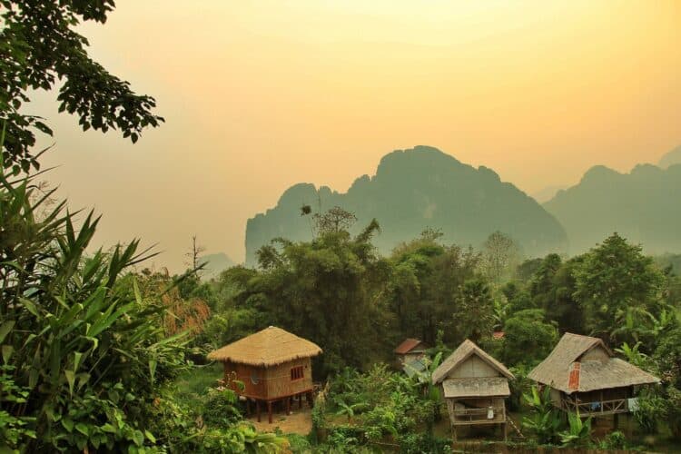 Village traditionnel au Laos avec maisons sur pilotis entourées de végétation tropicale et montagnes karstiques